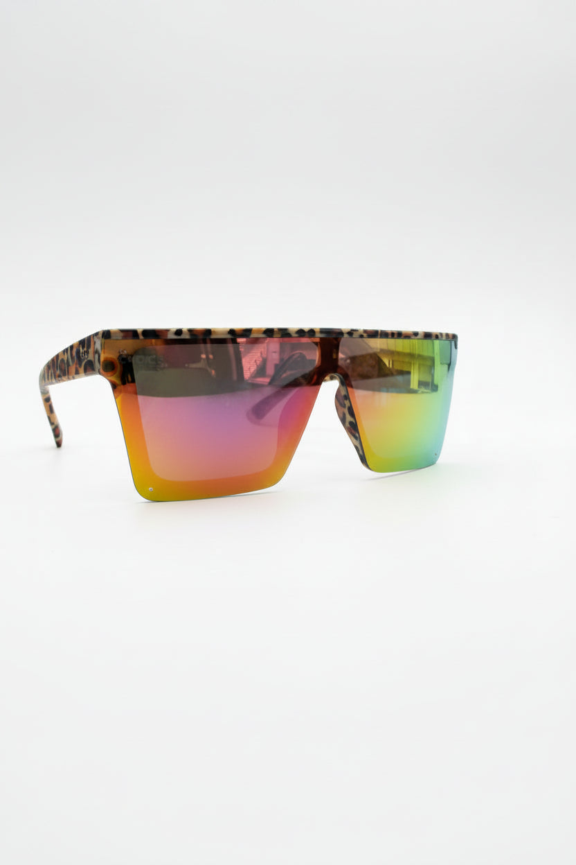 A pair of shield-style sunglasses with tortoiseshell-patterned frames and mirrored lenses showing a green-to-yellow gradient, resting on a black leather surface with a branded pop-up canopy in the background.