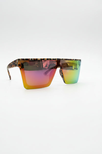 A pair of shield-style sunglasses with tortoiseshell-patterned frames and mirrored lenses showing a green-to-yellow gradient, resting on a black leather surface with a branded pop-up canopy in the background.